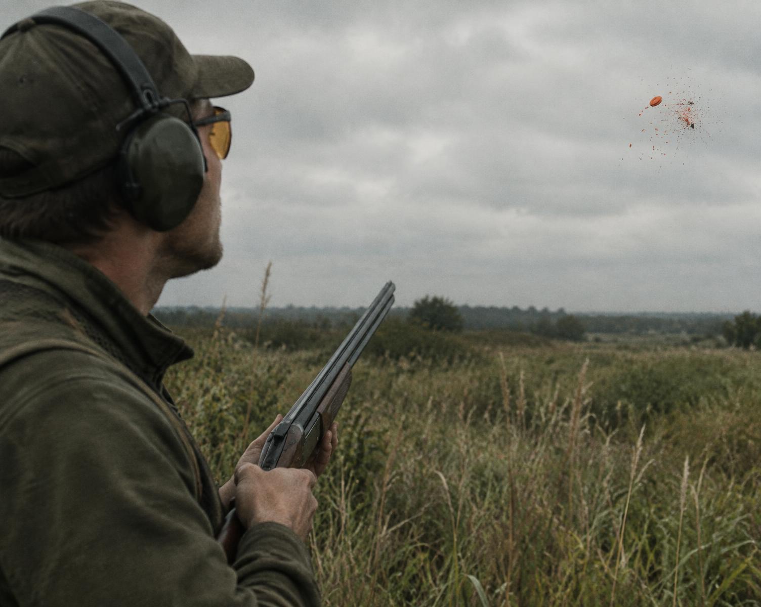 Competitor in ear protection and shooting glasses watching a clay target break over a grass field