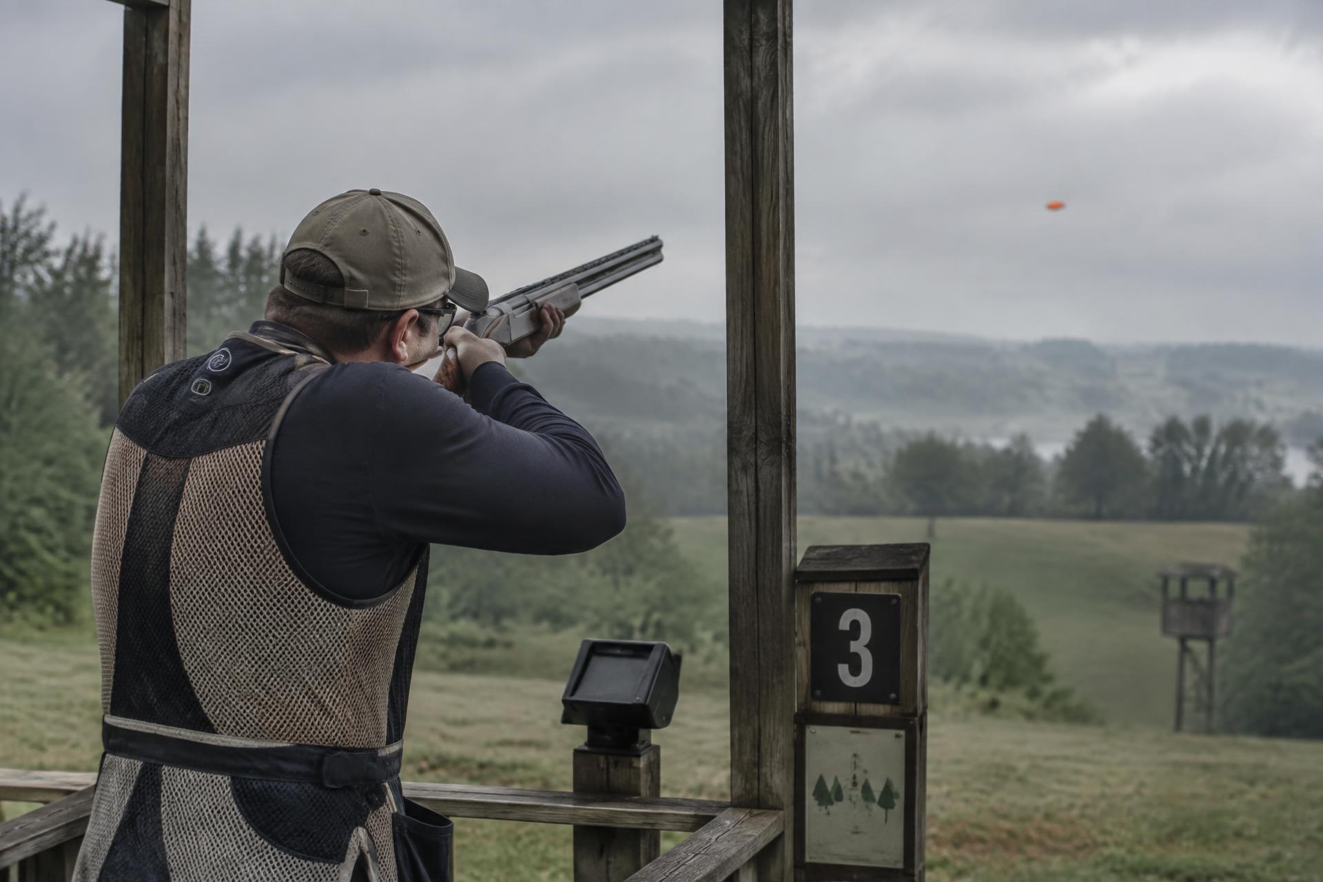 Sporting clays competitor mounted on a shotgun at a station, orange clay target in flight over a misty range